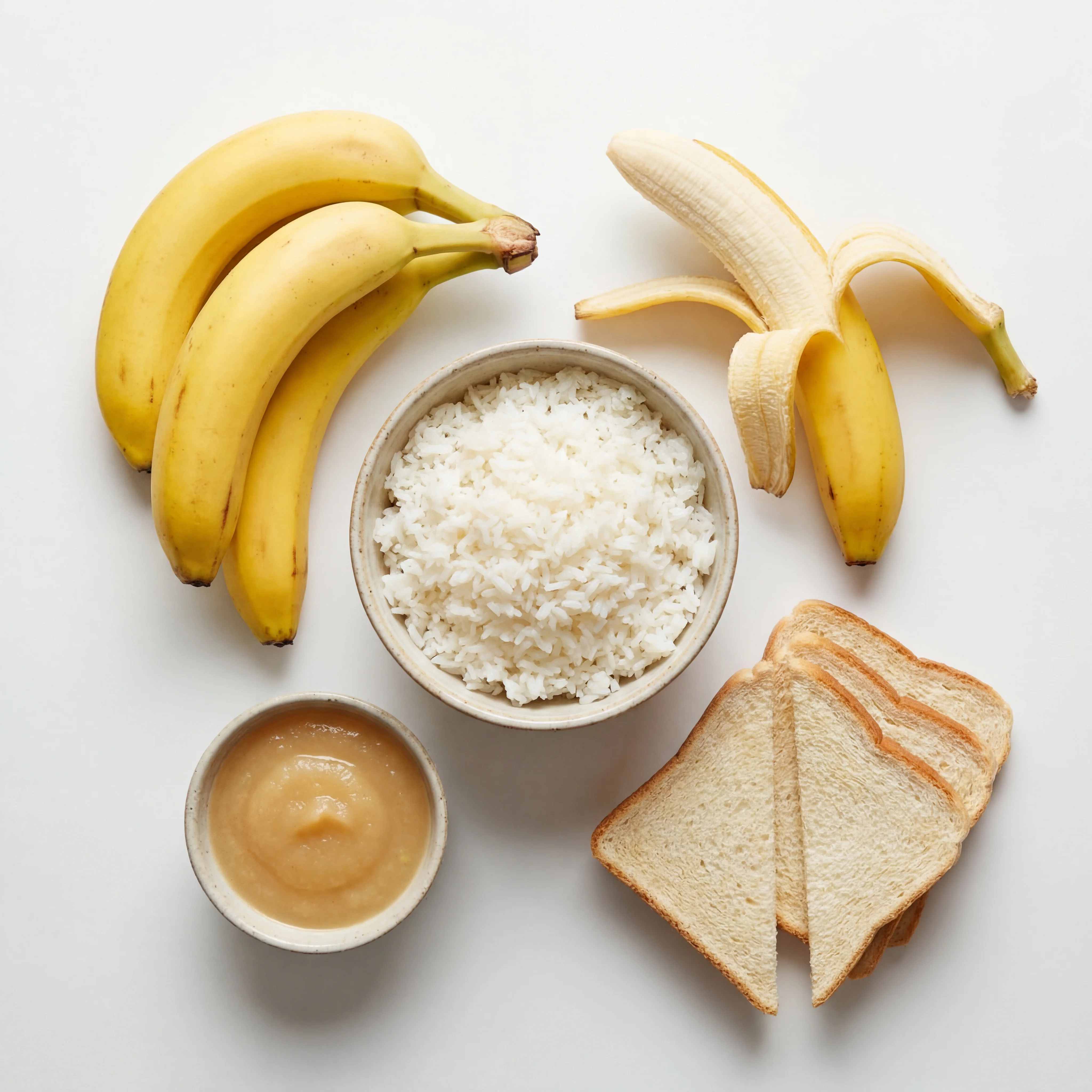 Arrangement of BRAT diet recovery foods including bananas, plain rice, applesauce, and dry toast on a light background